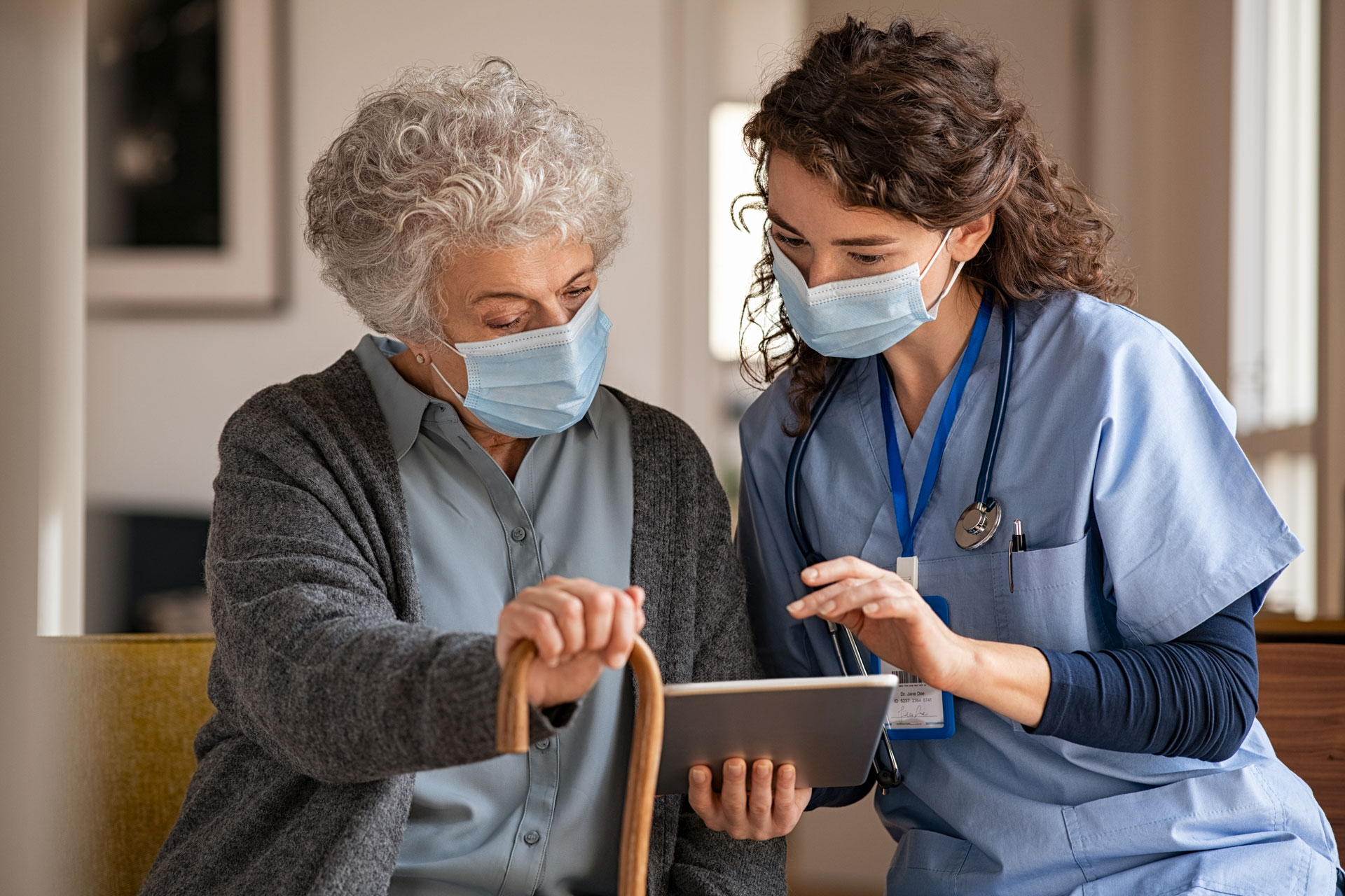 iStock-1284869080 Young doctor and senior woman going through medical record on digital tablet during home visit wearing face protective mask.
